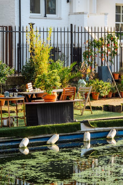 A garden situated beside a canal features a neatly maintained lawn with short, lush grass bordered by a narrow paved pathway. There are various potted plants, including small shrubs and flowering plan