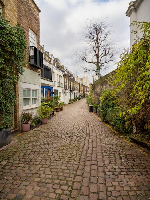 A narrow cobblestone street in Limehouse, lined with charming terraced houses featuring small front gardens with potted plants and shrubs. On the left, white and pastel-colored facades with windows an