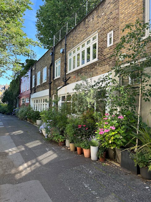A narrow cobblestone street in Limehouse, lined with charming terraced houses featuring small front gardens with potted plants and shrubs. On the left, white and pastel-colored facades with windows an